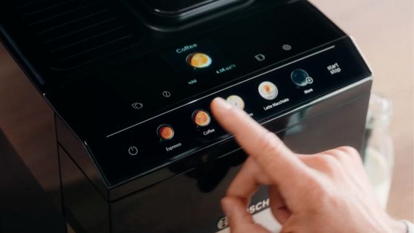 Un doigt touche le bouton "Café" sur le panneau de commande d'une machine à café Bosch, qui affiche des icônes pour différentes options de café, dont "Espresso", "Café", "Cappuccino", et "Latte Macchiato". La machine a une finition noire et moderne, et un logo Bosch est visible en bas.