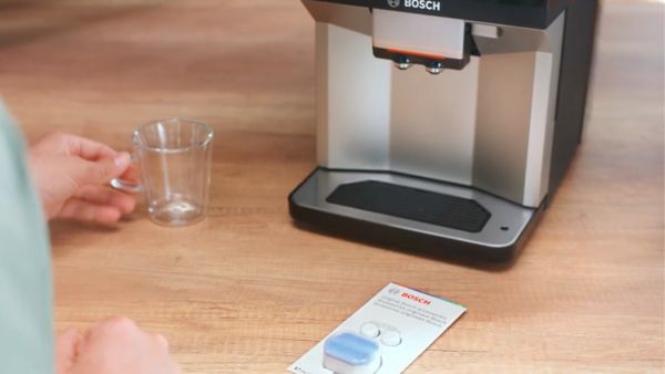 Une personne place une tasse en verre sur le plateau d'une machine à café Bosch. À côté, un paquet de pastilles Bosch est visible, contenant deux pastilles bleues, le tout sur une table en bois clair.