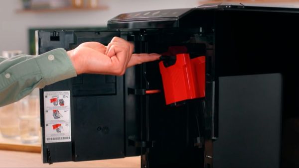 Une personne pointe du doigt un composant rouge à l'intérieur d'une machine à café Bosch, dont la porte est ouverte. Des instructions sont affichées sur le côté, et l'arrière-plan montre une cuisine avec des éléments en bois.