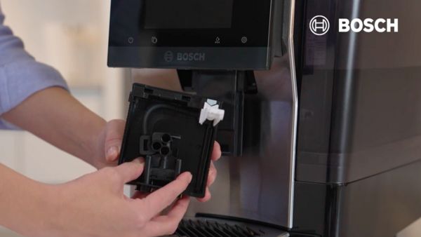 Une main tient un composant noir, probablement un filtre ou un réservoir, devant une machine à café Bosch. L'écran tactile de la machine est visible au-dessus, avec le logo Bosch en haut à droite de l'image.