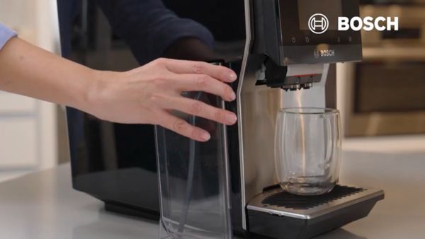 Une main retire un réservoir transparent d'une machine à café Bosch, pendant qu'un verre est placé sur le plateau en dessous. La machine est moderne et élégante, avec un logo Bosch visible en haut à droite de l'image.
