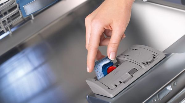 A hand placing a dishwasher tablet in the detergent compartment of the dishwasher.