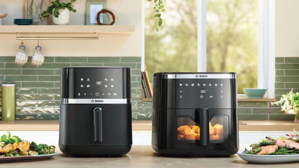 Ένας ξύλινος πάγκος κουζίνας, με πράσινες αποχρώσεις σε πλακάκια, φυτά αλλά κι φαγητά και πάνω του δύο Air Fryers σε διαφορετικά μεγεθη.