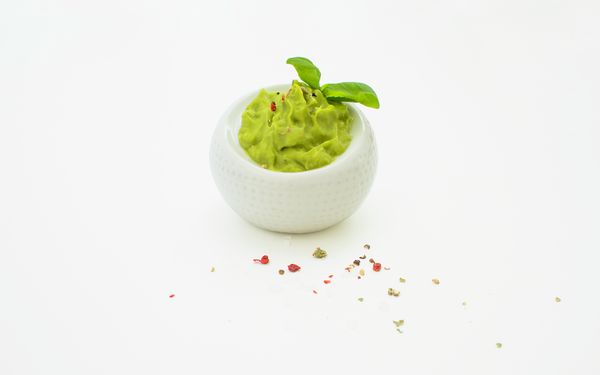 A half-cut avocado next to pepper, garlic and a prepared guacamole dip with a piece of lime.
