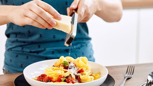 Eine stehende Person hobelt frischen Parmesan über ein Nudelgericht in einem weißen Teller mit schwarzem Unterteller auf einem Holztisch.