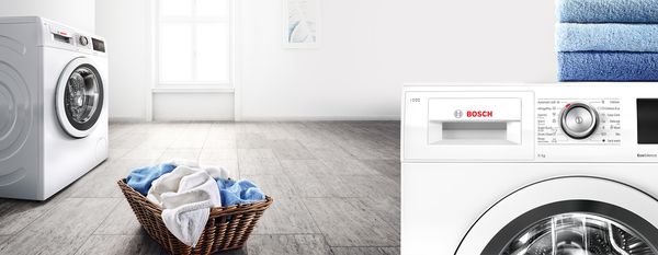 Folded towels resting on top of Bosch Washing machine with full washing hamper on floor.