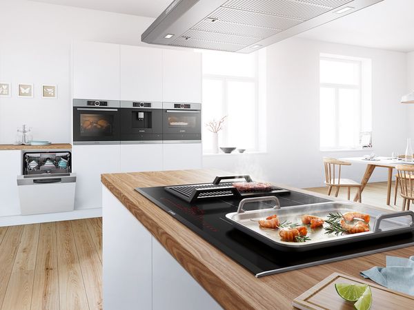 Modern white kitchen with black Bosch oven, dishwasher, hob, on which meat and prawns are cooking.