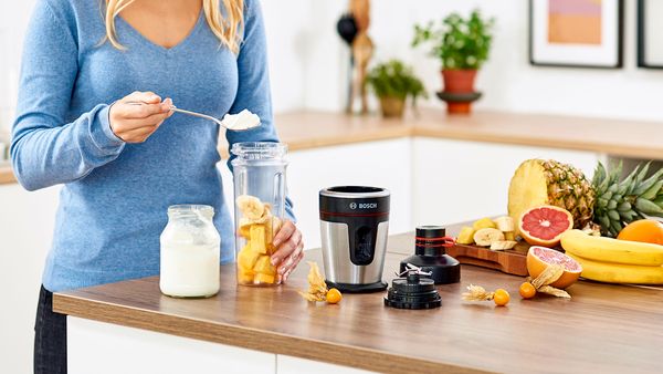 Eine Frau bereitet an der Kücheninsel einen Smoothie zu. Sie gibt Joghurt und frisch aufgeschnittenes Obst in ein Gefäß, um alles mit dem VitaPower Serie 2 Mini-Standmixer zu einem Smoothie zu verarbeiten.
