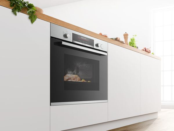 Bosch ovn med kødret indeni, indbygget i hvid køkkenø med træbordplader. Grøntsager ligger ovenpå.