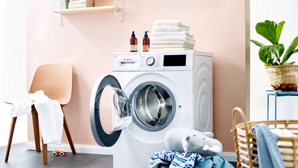  Eine Waschmaschine mit geöffneter Trommeltür steht in einer Waschküche, links daneben ein Stuhl. Auf dem Boden und Stuhl liegen Schmutzwäsche und ein Nashorn-Stofftier.