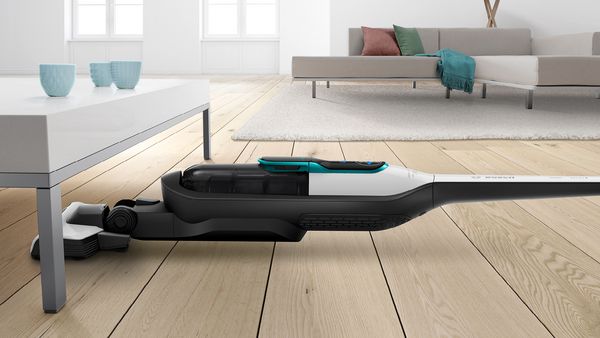 A cordless vacuum cleans under a living room table. There is a couch in the background.