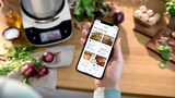 Eine Hand hält ein Smartphone mit der Home Connect App, die Rezeptvorschläge zeigt, während die Bosch Cookit Küchenmaschine, die kocht, im Hintergrund steht.