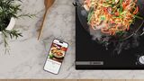 Vue de dessus d'un smartphone posé à côté de la plaque de cuisson dont l'écran affiche l'application BetterFood de Bosch.