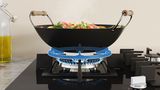 Un wok est posé sur la plaque de cuisson au gaz. Sous le wok se trouve un anneau de wok qui s'allume en bleu.