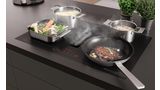 Strak keukenblad met inductiekookplaat, sissende steak, stoomende groenten, pasta en champignons in pannen.