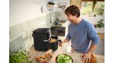 A young man slides a drawer filled with raw food into his Bosch Air Fryer, preparing for cooking in a bright kitchen.