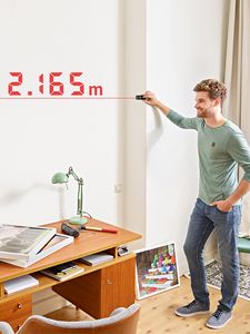 Ein Mann steht misst die Wand hintern einem Schreibtisch mit einem Laser-Entfernungsmesser. Die Distanz von 2.165 m steht and der Wand.