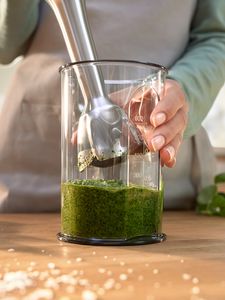 An immersion blender hovers over a beaker full of freshly made green pesto standing on a wooden counter.