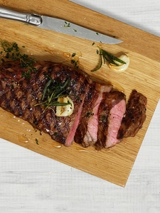 A piece of beef roast resting on a wooden chopping board with a knife and herbs.