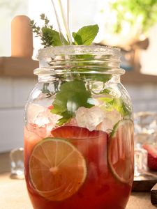 Borcan din sticlă cu suc proaspăt preparat cu accesoriul pentru stors citrice, cu cuburi de gheaţă şi felii de fructe proaspete, pe un blat de bucătărie.