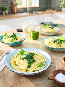 Fyra skålar pasta med grön pesto arrangerade på ett köksbord med en burk pesto i mitten.