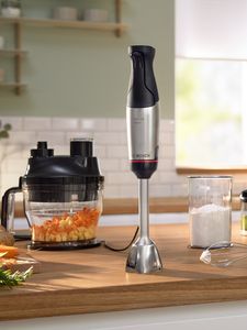 A woman smiling in a modern kitchen while using a Bosch blender. Fresh ingredients are visible in a bowl next to her.