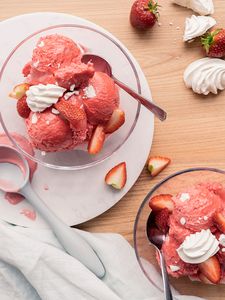 Två dessertglas med jordgubbsglass, garnerade med jordgubbar och maräng på en träyta.