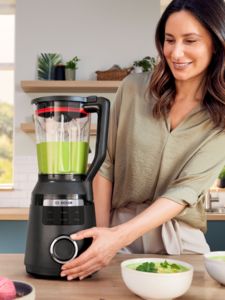 Uma mulher a sorrir numa cozinha moderna enquanto utiliza uma liquidificadora Bosch. Os ingredientes frescos estão visíveis numa tigela ao lado dela.