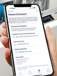 Dłoń trzymająca telefon przed zmywarką, z aplikacją pytającą, czy klient wymaga pomocy od Działu Obsługi Klienta.
