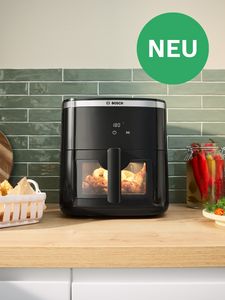 Bosch Heißluftfritteuse auf Küchenarbeitsplatte, kocht Huhn, mit 'NEU' Label im grünen Kreis.