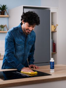 Uma pessoa em uma camisa de ganga limpa uma placa em vidro preta com um pano amarelo, enquanto uma embalagem do produto de limpeza aparece junto á placa, numa cozinha limpa e moderna. 