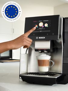 Κάποιος πιέζει την οθόνη αφής στη μηχανή καφέ VeroCafe Σειρά 8 στον πάγκο μιας κουζίνας με ένα φλιτζάνι cappuccino και μια σημαία της Ευρωπαϊκής Ένωσης στην πάνω γωνία της εικόνας.