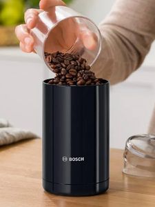 Una persona versa chicchi di caffè in un macinacaffè Bosch nero, su un tavolo di legno, in cucina. In secondo piano, un panno grigio