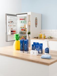 Uma cozinha brilhante com um frigorífico aberto, a mostrar a comida e uma bancada com produtos de limpeza, uma garrafa de spray de produto de limpeza e um pano azul.  