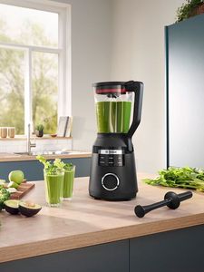 Una batdiroa de vaso Bosch plateada sobre una encimera de cocina, llena de un batido verde, rodeada de frutos secos y frutas.