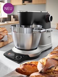 A sleek Bosch kitchen mixer with a stainless steel bowl sits on a countertop beside freshly baked bread, labeled "NEU" in purple.
