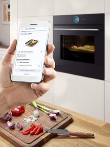 Smartfon sterujący wbudowanym piekarnikiem z warzywami na blacie w nowoczesnej kuchni.