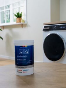 Uma mesa redonda de madeira exibe um produto descalcificador com embalagem azul com uma máquina de lavar roupa e plantas.