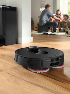 Ein Saugroboter von Bosch fährt über einen hellen Holzboden und wischt den Boden gründlich, während im Hintergrund eine Familie am Tisch sitzt und Zeit miteinander verbringt.