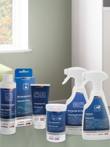 Uma variedade de produtos de limpeza, incluindo sprays e pastilhas, exibidos num fundo com uma parede verde perto de uma janela.