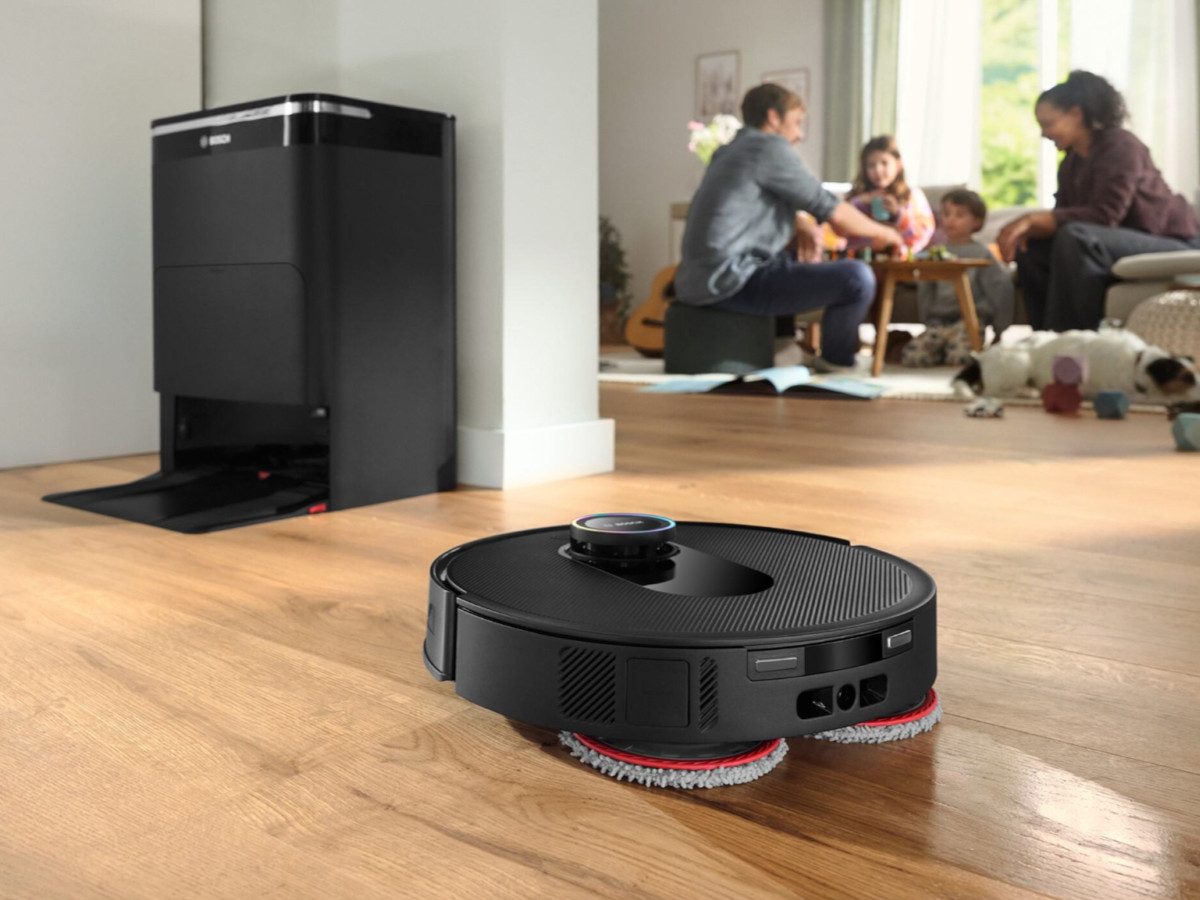 Schwarzer Saugroboter mit Wischpads reinigt einen Holzboden in einem Wohnzimmer, mit einer Familie und einem Hund im Hintergrund.
