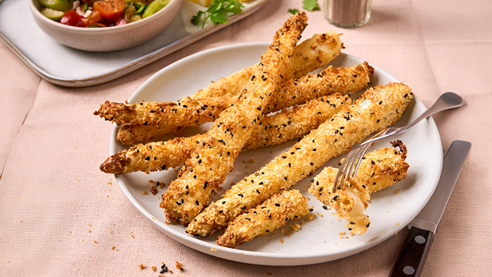 Knusprig gebackene Grissini mit Sesam auf einem wei&szlig;en Teller, daneben liegen Gabel und Messer, im Hintergrund steht eine Salatsch&uuml;ssel.