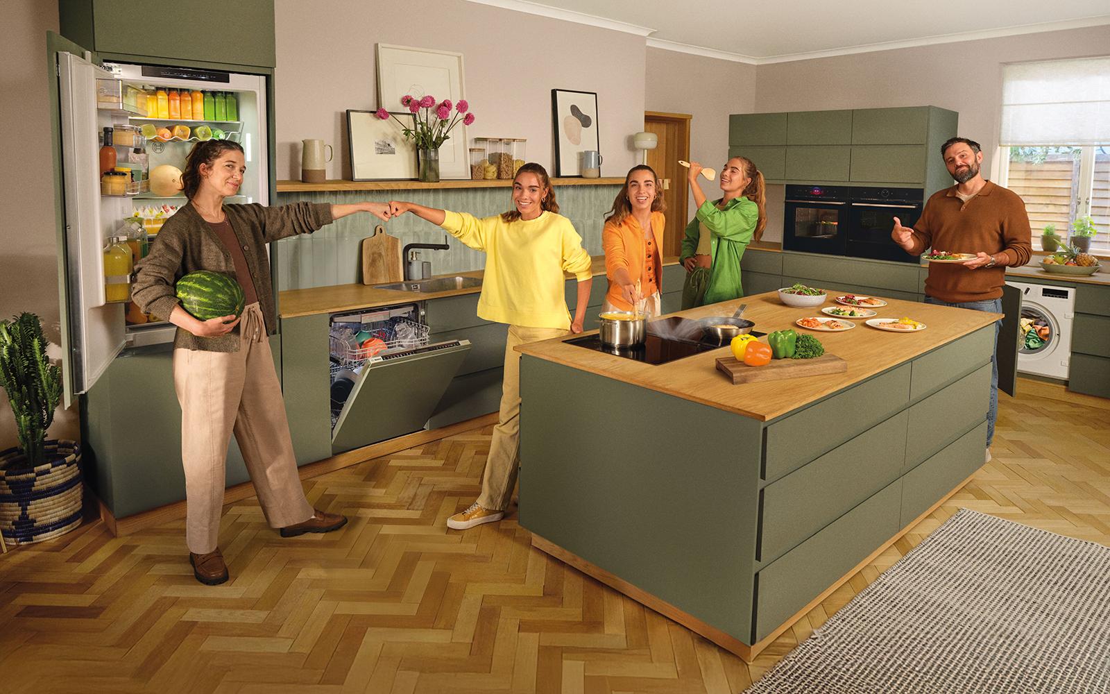 Cocina moderna con isla y electrodom&eacute;sticos Bosch (frigor&iacute;fico, lavavajillas, hornos, lavadora); cinco personas cocinando y compartiendo.