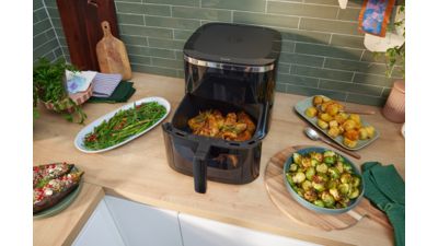 Friggitrice ad aria aperta su un piano cucina con pollo nel cestello, circondata da contorni di verdure e patate arrosto