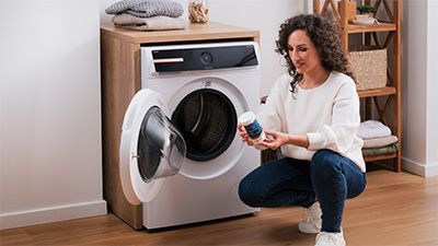 Eine Person kniet neben einer Waschmaschine und h&auml;lt einen Beh&auml;lter mit Waschmaschinenreiniger in der Hand. In der Umgebung befinden sich ein Holzregal und ordentlich gefaltete Handt&uuml;cher. 