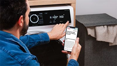 Ein Mann nimmt Einstellungen an einer modernen Waschmaschine vor, w&auml;hrend er die Anleitungen auf seinem Smartphone nachliest. 