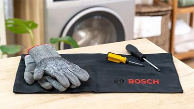 Eine Werkzeugunterlage der Marke Bosch liegt auf einer Holzoberfl&auml;che. Darauf befinden sich graue Arbeitshandschuhe, ein gelber Schraubendreher und ein schwarzer Schraubendreher. 