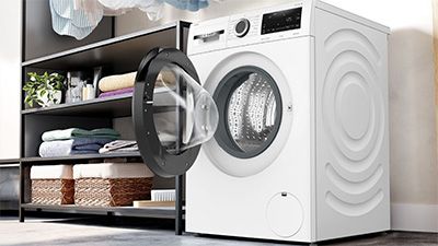 Eine wei&szlig;e Bosch Waschmaschine mit ge&ouml;ffneter T&uuml;r, die auf Probleme beim Waschen hindeutet. Daneben befinden sich ordentlich gefaltete Handt&uuml;cher und K&ouml;rbe auf einem Regal. 