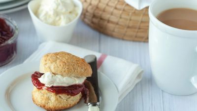 A freshly baked scone topped with cream and berry jam on a plate, with a cup of tea to the side