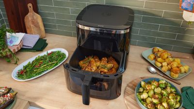 Friggitrice ad aria Bosch con cestello aperto e pollo arrosto all&rsquo;interno, circondata da contorni di patate, cavoletti e asparagi